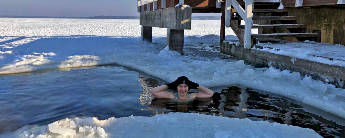 Na zdjęciu widać kobietę w czapce i rękawiczkach, morsującą w zimnej wodzie Jeziora Zegrzyńskiego, obok niej molo. W tle rozciąga się skuta lodem tafla Jeziora Zegrzyńskiego