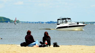 Na zdjęciu kobiety wypoczywające nad wodą na plaży, w tle płynie duża łódź motorowa