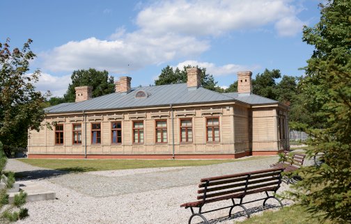 fot. Filia "Piaski" Muzeum Historycznego w Legionowie, https://www.facebook.com/people/Filia-Piaski-Muzeum-Historycznego-w-Legionowie/100063549297127/. Na zdjęciu widok z zewnątrz na filię "Piaski" Muzeum Historycznego w Legionowie, obok ławki do siedzenia