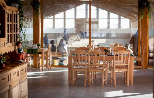 fot. stajniaojcowizna.pl/Restaurant. Na zdjęciu restauracja Stajnia Ojcowizna, stoliki z krzesełkami, z tyłu osoby jeżdżące na koniach