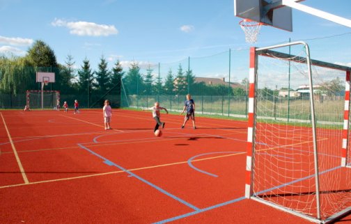 fot. www.nieporet.pl/sport-i-turystyka/obiekty-sportowe/. Na zdjęciu boisko wielofunkcyjne do gry w piłkę nożną, koszykową, dzieci grają na boisku, chłopiec uderza piłkę nogą w kierunku bramki, w tle drzewa, budynki mieszkalne