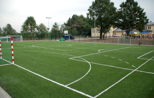 fot. www.nieporet.pl/sport-i-turystyka/obiekty-sportowe/#sportBoiska. Na zdjęciu boisko o nawierzchni sztucznej do gry w piłkę ręczną, koszykową, nożną, w tle budynek szkoły, przed budynkiem szkoły uczniowie