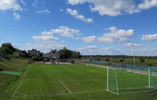 fot. wieliszewsp.szkolnastrona.pl/. Na zdjęciu boisko do gry w piłkę nożną, w tle plac zabaw dla dzieci, po prawej stronie Jezioro