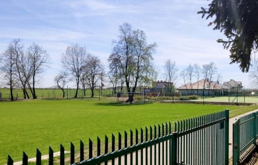 fot. sis.serock.pl/1471,boisko. Na zdjęciu boisko wielofunkcyjne, trawiaste w Jadwisinie, w tle duża bramka do piłki nożnej, domy, wzdłuż boiska po prawej stronie zielone ogrodzenie
