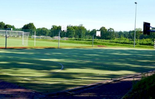 fot. sis.serock.pl/3926,boisko. Na zdjęciu boisko do gry w koszykówkę i piłkę nożną o sztucznej nawierzchni, w tle boisko  do piłki nożnej, w tle z tyłu bramka do piłki nożnej, drzewa