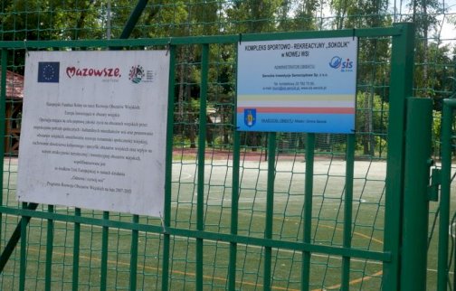 fot. sis.serock.pl/3836,nowa-wies. Na zdjęciu tablice wiszące na ogrodzeniu od boiska wielofunkcyjnego w Nowej Wsi - Kompleks Sportowo - Rekreacyjny "Sokolik" w Nowej Wsi, na białej tablicy po lewej stronie zdjęcia flaga Unii Europejskiej, napis MAZOWSZE serce Polski, za ogrodzeniem boisko wielofunkcyjne ze sztuczną nawierzchnią w Nowej Wsi