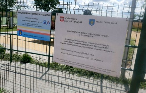 fot. sis.serock.pl/3838,gasiorowo. Na zdjęciu na ogrodzeniu dwie tablice. Jedna z napisem KOMPLEKS SPORTOWO - REKREACYJNY GĄSIOROWO, MODERNIZACJA BOISKA WIELOFUNKCYJNEGO W GĄSIOROWIE, w tle za ogrodzeniem siłownia zewnętrzna w plenerze, plac zabaw