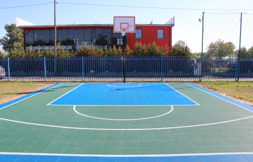 fot. https://www.osirjablonna.pl/baza-sportowa/boisko-w-suchocinie. Na zdjęciu boisko o sztucznej nawierzchni do gry w piłkę koszykową, w tle z tyłu budynek