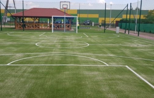 fot. sis.serock.pl/3839,stasi-las. Na zdjęciu boisko o sztucznej nawierzchni do gry w piłkę nożną oraz koszykową, w tle drewniana altana, ściana energochłonna od drogi samochodowej