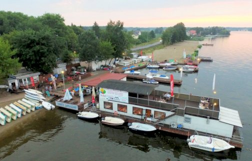 fot. www.water-club.pl. Na zdjęciu barka - restauracja, łodzie, rowerki wodne ułożone na brzegu, z tyłu plaża, akwen Jeziora Zegrzyńskiego