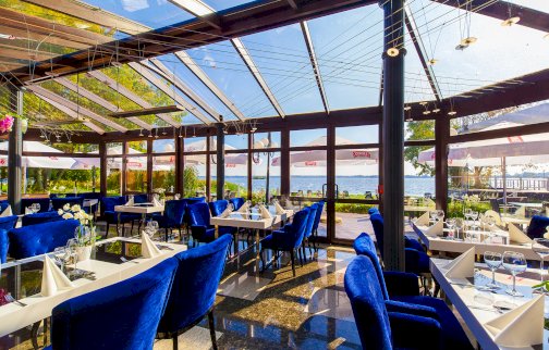 fot. www.hotel500.com.pl. Na zdjęciu Restauracja w Zegrzu Hotel 500, zastawa na stołach, kieliszki, niebieskie fotele, złożone serwetki, przeszklone okna restauracji, na zewnątrz parasole, taras, leżaki, widok na Jezioro Zegrzyńskie