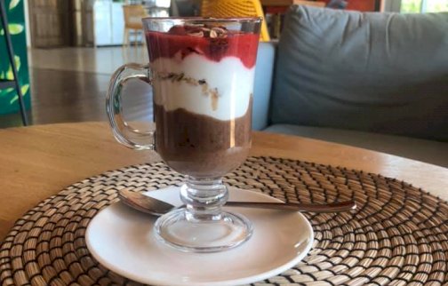 fot. https://www.facebook.com/PasjaZycia. Na zdjęciu deser z musem w szklance, talerzyk, łyżeczka  do jedzenia, w tle biblioteczka z książkami na półkach, fragment sofy do siedzenia, w tle widoczne drzwi wejściowe do kawiarni