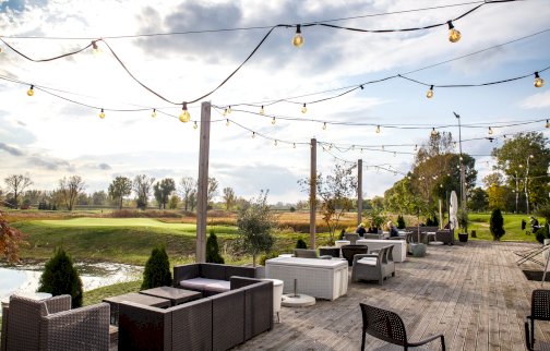 fot. firstwarsaw.pl. Na zdjęciu taras restauracji First Warsaw Golf ze stolikami i fotelami, w tle pole golfowe z dołkami