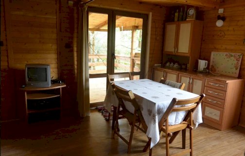 fot. wwww.swiecienica.wixsite.com/rybaczowka. Na zdjęciu drewniany stół z krzesłami, mały telewizor na szafce, czajnik elektryczny, zegar, drzwi balkonowe, za oknem taras