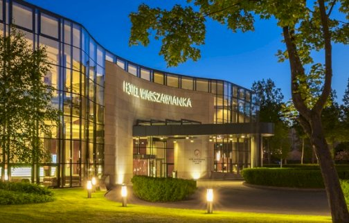 fot. www.warszawianka.pl. Na zdjęciu Hotel Warszawianka z zewnątrz, oświetlone pomieszczenia wewnątrz budynku, oświetlone alejki, drzewa