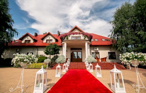 fot. www.magnacka.pl. Na zdjęciu hotel sala gościnna magnacka, długi, czerwony dywan przed wejściem, przy wejściu kwiaty, białe lampiony rozstawione wzdłuż dywanu