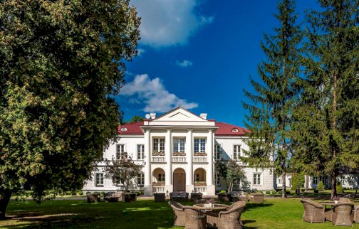 Fot. www.palac-zegrzynski.pl. Na zdjęciu widok na Hotel Pałacu Zegrzyńskiego w Zegrzu, wiklinowe krzesełka ze stolikami leżące na trawie, zielony trawnik z drzewami