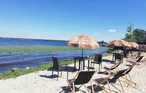 fot. www.magnacka.pl. Na zdjęciu piaszczysta plaża, na niej leżaki do opalania się, stoliki z krzesełkami, w tle Jezioro Zegrzyńskie