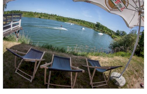 fot. plazajablonna.pl. Na zdjęciu trzy leżaki nad wodą, nad nimi parasol, z tyłu osoba płynie na desce, wakeboardzie na wodzie