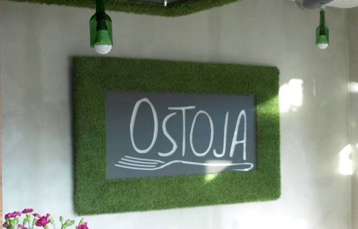 fot. Ostoja, https://www.facebook.com/p/Ostoja-100046406899706/?locale=pl_PL.  Na zdjęciu szyld, na szyldzie napis OSTOJA oraz grafika widelca, stolik z krzesłami, kwiat