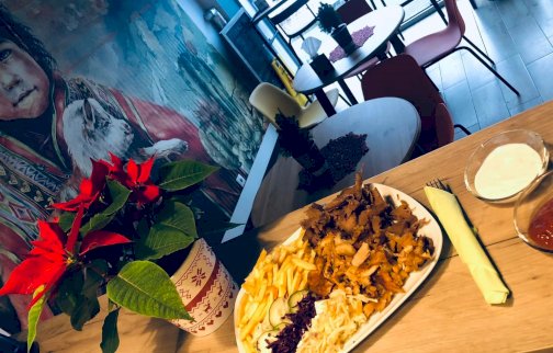 fot. facebook Kebabownia, https://www.facebook.com/p/Kebabownia-100063488023501/?locale=pl_PL. Na zdjęciu restauracja Kebabownia Wieliszew (Osiedle Royal), danie z siekanym mięsem, frytkami i surówkami, na stole sztućce, sosy: pomidorowy, czosnkowy, kwiat w doniczce, stoliki z krzesłami w restauracji, tapeta chłopca na ścianie