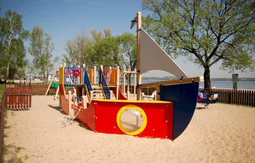 fot. www.nieporet.pl. Na zdjęciu plac zabaw przy plaży w Nieporęcie, urządzenie do zabawy  w kształcie dużego statku, teren placu zabaw posiada ogrodzenie