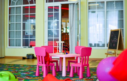 fot. www.windsorhotel.pl. Na zdjęciu mały stolik z krzesełkami do siedzenia dla dzieci, z tyłu drzwi wejściowe do sali zabaw, po obu stronach drzwi okna, obok stolika obustronna tablica do rysowania, duża piłka do ćwiczeń