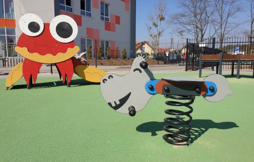 fot. https://www.osirjablonna.pl/rekreacja/place-zabaw. Na zdjęciu urządzenia zabawowe dla dzieci: bujak sprężynowy, krab, z tyłu fragment budynek przedszkola gminnego w Chotomowie