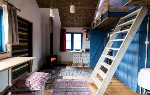 fot. Kurnik, https://siedliskoleluja.pl/nocleg/#!/kurnik. Na zdjęciu kominek, drewniane schody prowadzące do łóżka piętrowego, dywany leżące na podłodze, poduszka na skrzynce, stolik, z tyłu okno, lampy pod sufitem, otwarte skrzydło drzwiowe w kolorze niebieskim po prawej stronie