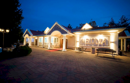 fot. https://salamelodia.pl/galeria/. Na zdjęciu sala bankietowa & pensjonat Melodia. Oświetlony budynek, stolik z ławkami