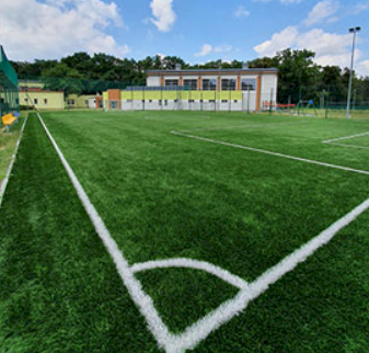 fot. www.spb.nieporet.pl. Na zdjęciu fragment boiska do piłki nożnej, wyznaczone linie na boisku, w tle Szkoła Podstawowa w Białobrzegach, oświetlenie boiska (słup oświetleniowy)