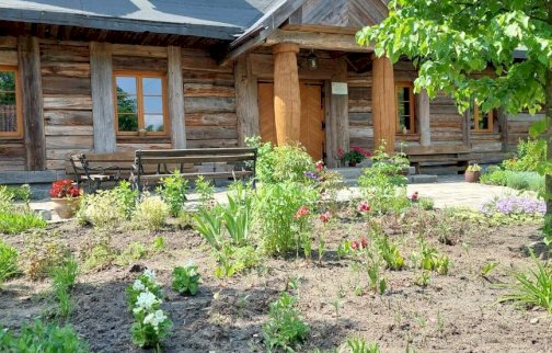 fot. facebook Skansen w Kuligowie nad Bugiem. Na zdjęciu widok na skansen w Kuligowie nad Bugiem w wiosennej aurze. Kwiaty rosnące przed skansenem, przy skansenie ławki do siedzenia