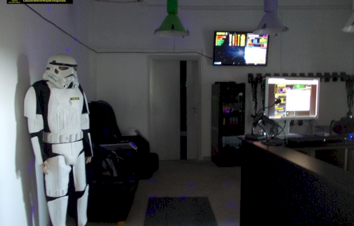 fot. https://www.laser-war.pl/jsp/main.jsf. Na zdjęciu postać bohatera filmowego, włączony wyświetlacz na telewizorze, włączony ekran monitora komputera, z tyłu przy ścianie lodówka, ciemne fotele do gry