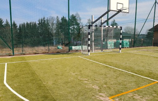 fot. https://serock.pl/serockie-inwestycje-samorzadowe/obiekty/wierzbica/boisko-wielofunkcyjne. Na zdjęciu fragment boiska z trawy syntetycznej - boisko (z tablicą do kosza) do piłki koszykowej oraz boisko do gry w piłkę nożną. Siatka ochronna z tyłu boiska, w tle z tyłu drzewa, domy mieszkalne jednorodzinne