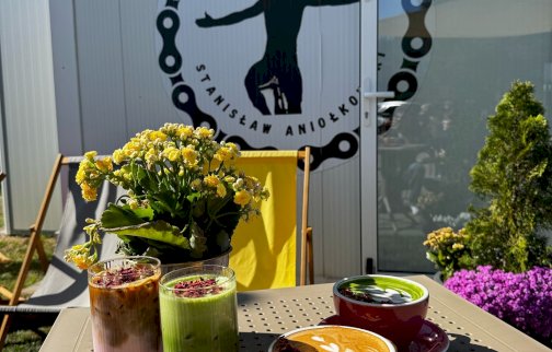 fot. facebook Port Kawiarnia, https://www.facebook.com/photo?fbid=562797863541016&set=pcb.562797903541012&locale=pl_PL. Na zdjęciu dwie filiżanki z kawą, z dodatkami, dwie wysokie szklanki z shakiem owocowym, obok kwiat doniczkowy na stole, z tyłu logo z napisem Port Kawiarnia Stanisław Aniołkowski. Leżaki rozłożone na trawie przed kawiarnią