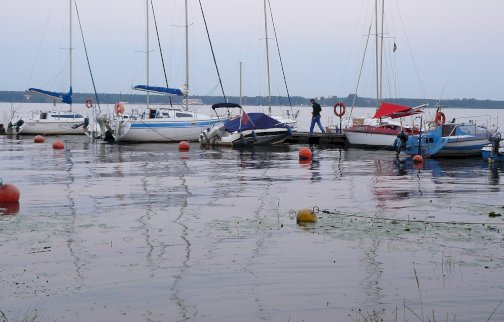 fot. Klub Dolomit w Jadwisinie. Na zdjęciu widać jachty zacumowane przy pomoście na spokojnej wodzie Zalewu Zegrzyńskiego w porcie Klubu Dolomit w Jadwisinie. Mężczyzna porusza się na pomoście