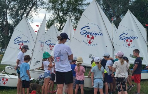 fot. Klub Sportowy "Spójnia-Warszawa". Grupa dzieci wraz z instruktorami stoi przy małych żaglówkach Optimist z żaglami klubu sportowego Spójnia - Warszawa