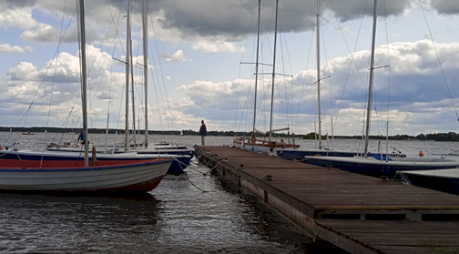 fot. Ośrodek MOS-2, filia w Zegrzu Południowym. Na fotografii drewniany pomost wychodzący na akwen, przy którym zacumowane są jachty żaglowe, a w tle rozciąga się szeroka tafla wody, z tyłu na pomoście stoi mężczyzna