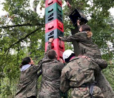 Fot. EVENT EXTREME FORT ROBSON. Grupa osób w strojach maskujących wspólnie buduje wysoką wieżę ze skrzynek, podając kolejne elementy osobie stojącej na jej szczycie