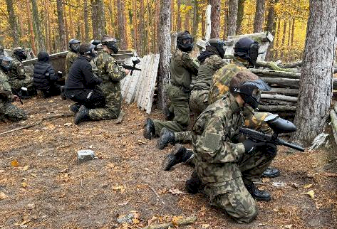 Fot. EVENT EXTREME FORT ROBSON. Grupa osób w strojach maskujących i maskach ochronnych klęczy w lesie za drewnianą przeszkodą i trzyma markery do paintballa