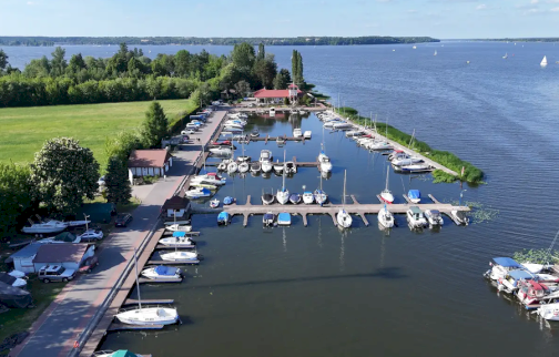 fot. TKKF Wodnik. Na zdjęciu przystań jachtowa z zacumowanymi łodziami i jachtami nad jeziorem, otoczoną zielenią