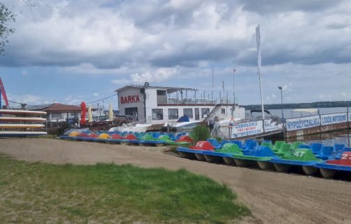 Fot. Water-Club Nieporęt. Na zdjęciu plaża, wypożyczalnia rowerków wodnych, kajaków, pomost, widoczny napis na pomoście: wypożyczalnia, łodzie motorowe bez patentu, restauracja BARKA, za barką Jezioro Zegrzyńśkie
