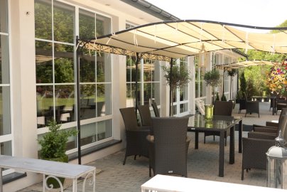 Powiększ obraz: fot. restauracjaoranzeria.com. Na zdjęciu zewnętrzny taras Restauracji Oranżeria, stoliki z krzesełkami, zadaszenie nad stolikami, kwiaty