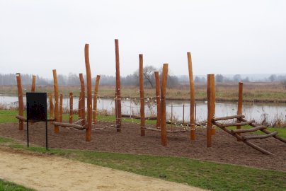 Powiększ obraz: fot. Krzysztof Kozań. Na zdjęciu drewniane słupy, zamocowana na słupach ścieżka do przechodzenia z linami, z tyłu w tle Jezioro Wieliszewskie, łąka