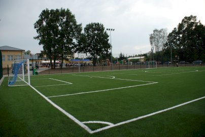 Powiększ obraz: fot. www.nieporet.pl/sport-i-turystyka/obiekty-sportowe/#sportBoiska.  Na zdjęciu boisko o nawierzchni sztucznej do gry w piłkę ręczną, koszykową, nożną, w tle budynek szkoły, przed budynkiem szkoły uczniowie