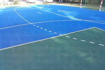 Powiększ obraz: fot. splajski.edupage.org/. Na  zdjęciu boisko ze sztucznej nawierzchni do gry w piłkę nożną, bramka do piłki nożnej w tle, z boku samochody na parkingu