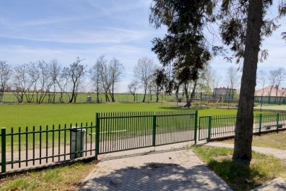 Powiększ obraz: fot. sis.serock.pl/1471,boisko. Na zdjęciu boisko wielofunkcyjne, trawiaste w Jadwisinie, w tle duża bramka do piłki nożnej, domy, wzdłuż boiska po prawej stronie zielone ogrodzenie