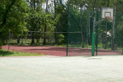 Powiększ obraz: fot. sis.serock.pl/3836,nowa-wies. Na zdjęciu boisko do gry w piłkę koszykową z koszem do rzucania piłek od koszykówki, w tle  boisko do gry w piłkę siatkową, drzewa