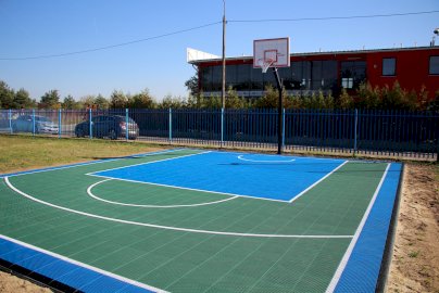 Powiększ obraz: fot. https://www.osirjablonna.pl/baza-sportowa/boisko-w-suchocinie. Na zdjęciu boisko o sztucznej nawierzchni do gry w piłkę koszykową, w tle z tyłu budynek, samochody zaparkowane na chodniku