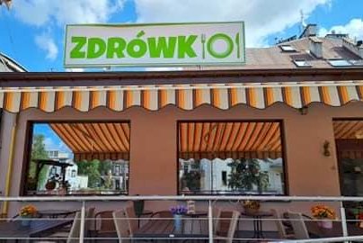 Powiększ obraz: fot. zdrowkolegionowo.org. Na zdjęciu restauracja ZDRÓWKO, taras - stoliki z krzesełkami do siedzenia, obszerne okna, w tle fragmenty budynków
